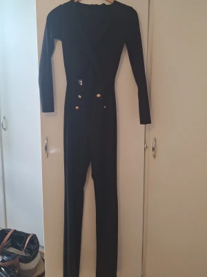 Svart långärmad jumpsuit med knappar - Snygg svart jumpsuit med långa ärmar och smal passform. Framsidan har dekorativa guldknappar som ger en cool touch. Djup v-ringning och hel längd på benen. Perfekt för dig som vill sticka ut med en stilren look.