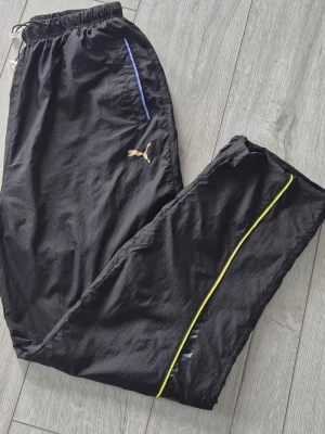 Black Puma Track Pants - These are black Puma track pants featuring a gold Puma logo on the left thigh. The pants have a blue accent on the pocket and a yellow stripe running down the side of the leg.