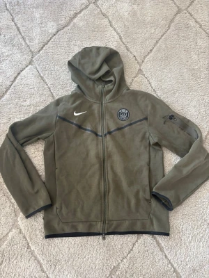 Olivgrön Nike PSG hoodie med dragkedja - Snygg olivgrön hoodie från Nike med PSG-logga på bröstet och hel dragkedja framtill. Huva och långa ärmar, diskreta svarta detaljer och fickor på sidorna. Perfekt för dig som gillar sportig stil och vill representera Paris Saint-Germain.