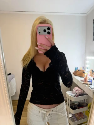 Svart spetsig långärmad topp - Säljer en svart långärmad topp i spets med v-ringad urringning och transparenta detaljer. Toppen är tight och har ett feminint, elegant uttryck med blommigt spetsmönster. Perfekt för dig som vill ha en trendig och snygg look.