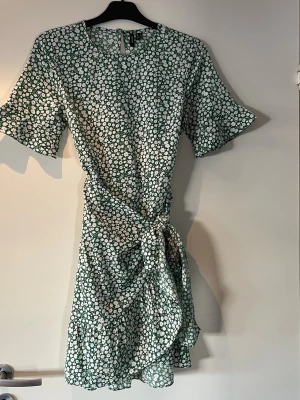 Grön blommig omlottklänning Vero Moda - Supersöt grön omlottklänning från Vero Moda med vitt blommönster. Klänningen har korta ärmar, volangkant och knyt i midjan för en snygg siluett. Perfekt för sommarens alla tillfällen!