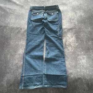 Blå Diesel vintagejeans för damer i storlek S - Säljer ett par blåa bootcut jeans med stora bakfickor med lock och knappar samt sidoficka på benet. Jeansen har klassisk denimlook med ljus kontrastsöm och vida ben. Perfekta för en avslappnad och trendig stil.