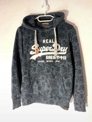 Blå mönstrad hoodie från Superdry - Snygg blå hoodie från Superdry med blommigt mönster och stor vit logga på bröstet. Hoodien har huva med breda gråa dragsnören och en klassisk känguruficka framtill. Perfekt för en avslappnad och trendig stil.