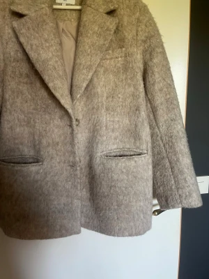 Beige ullkappa med klassisk krage från NAKD - Stilren beige kappa i ullmix med klassisk krage och två framfickor. Kappan har rak passform och dold knäppning framtill. Perfekt för kyliga dagar och ger en tidlös look.