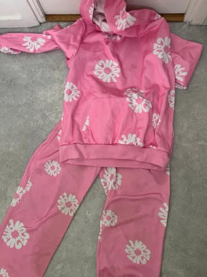 Pink Hoodie and Sweatpants Set with White Flower Pattern - This is a matching set consisting of a pink hoodie and sweatpants. Both pieces feature a repeating white flower pattern. The hoodie has a front pocket and a hood, while the sweatpants have an elastic waistband and side pockets. The set appears to have some visible stains or discoloration on both the hoodie and pants.
