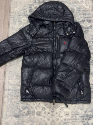 Svart dunjacka från Polo Ralph Lauren - Snygg svart dunjacka från Polo Ralph Lauren med röd logga på bröstet. Jackan har huva, dragkedja framtill och två sidofickor med dragkedja. Perfekt för kalla vinterdagar och riktigt skön att bära. 