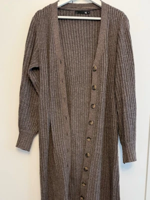 Lång ribbstickad cardigan  - Mjuk och skön långkofta från Lager 157 i en fin brungrå/mullvadsfärgad nyans. Koftan är ribbstickad med hel knapprad framtill och långa ärmar. Perfekt att bära både öppen och stängd under kyligare dagar.