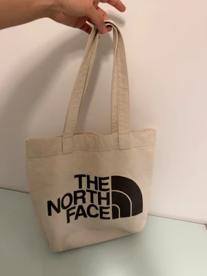 Beige shoppingväska The North Face - Snygg beige shoppingväska från The North Face i slitstark canvas. Väskan har två långa handtag och stor logga i svart på framsidan samt en mindre logga på baksidan. Perfekt för att bära med sig allt man behöver till skolan eller stan.