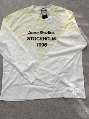 Vit långärmad topp Acne Studios - Vit långärmad topp från Acne Studios med tryck 'STOCKHOLM 1996' på bröstet. Tillverkad i mjuk bomull med rund hals och enkel, clean design. Perfekt för dig som gillar minimalistisk stil och vill ha något från ett ikoniskt svenskt märke.