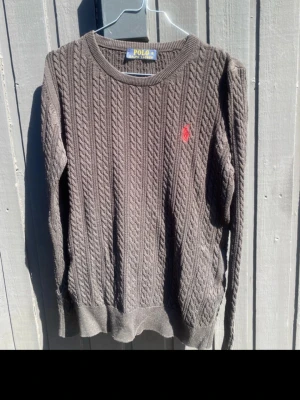 Grå kabelstickad tröja från  ralph lauren  - Kabelstickad grå tröja från Polo med rund halsringning och röd broderad logga på bröstet. Tröjan har ribbade muddar vid ärmslut och nederkant, och är tillverkad i ett mjukt stickat material. Perfekt för lager-på-lager-stil. Storlek Xl men liten i storleken så snarare en L-M