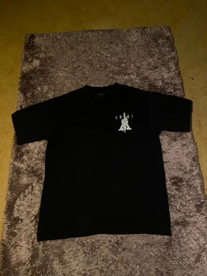 Svart Amiri t-shirt med tryck - Säljer en svart t-shirt från Amiri med stort grafiskt tryck på ryggen och mindre tryck på bröstet. T-shirten har klassisk passform och korta ärmar. Perfekt för dig som gillar streetwear och vill sticka ut med en unik design.
