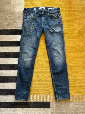 Blå slitna jeans med hål - Säljer ett par blå jeans med slitningar och hål på låren. Jeansen har klassisk femficksmodell, normal midja och raka ben. Snygg tvätt med ljusare partier och råa detaljer för en trendig look.