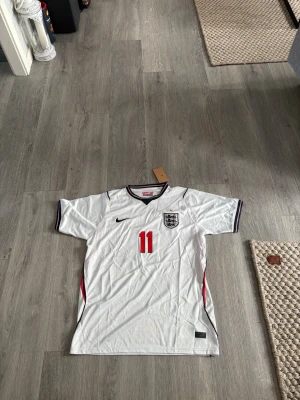 Vit England fotbollströja nr 11 - Säljer en vit England fotbollströja med rött nummer 11 på framsidan och klubbmärke på bröstet. Tröjan har svarta och röda detaljer vid ärmslut och hals. Perfekt för dig som älskar fotboll och vill stötta England.