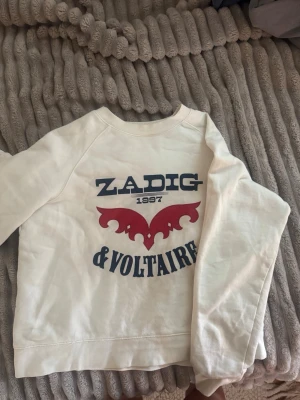 Vit sweatshirt från Zadig & Voltaire - Vit sweatshirt från Zadig & Voltaire med tryck i blått och rött framtill. Tröjan har rund halsringning och långa ärmar. Materialet känns mjukt och bekvämt, perfekt för en avslappnad stil. Snygg logga och coolt motiv på bröstet.