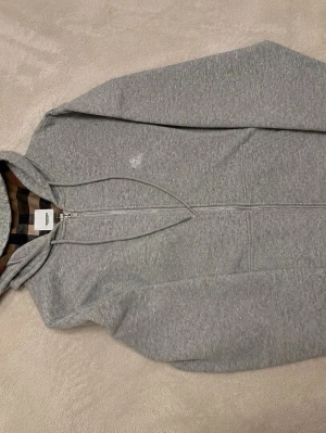 Grå zip hoodie med huva - Snygg gråmelerad hoodie med dragkedja och huva. Klassisk passform med dragsko i huvan och fickor framtill. Insidan av huvan har ett rutigt mönster som ger en extra detalj. Perfekt till chill eller streetwear-stil.