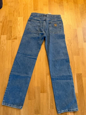 Blå raka jeans från Carhartt bootcut - Säljer ett par klassiska blå jeans från Carhartt med raka ben och hög midja. Jeansen har två bakfickor, varav en med Carhartt-logga i läder, samt bälteshällor. Tillverkade i slitstark denim som passar perfekt till en avslappnad stil. W30 L34