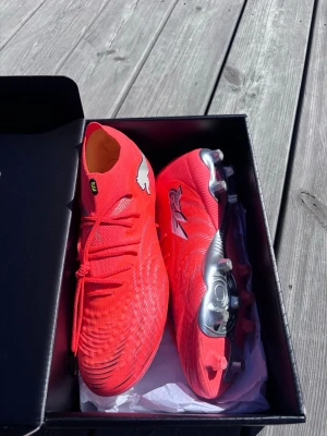 Helt Nya Puma Future - Helt nya fotbollsskor med hög kr och snörning. Skorna har en modern design med silvriga detaljer på sulan. Tillverkade i syntetmaterial för lätt vikt och bra passform. Perfekta för dig som vill sticka ut på fotbollsplanen.