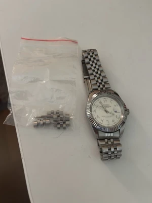  Seiko Klassisk silverfärgad armbandsklocka - Stilren armbandsklocka i silver med vit urtavla och metallarmband. Klockan har tydliga siffror och datumvisning. Extra länkar till armbandet medföljer så du kan justera storleken enkelt. Perfekt accessoar för dig som gillar en clean look.