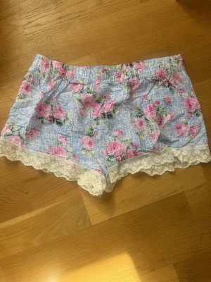Floral Lace-Trim Pajama Shorts - These are light blue pajama shorts with a pink rose floral print and white lace trim at the hem. The waistband is elasticated for comfort. The tag indicates the brand 'Love & Other Things' and the size as XS.