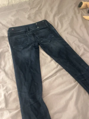 Mörkblå jeans med nitar - Snygga mörkblå jeans med smal passform och coola nitar på bakfickorna. Jeansen har slitningar och detaljer i silver på linningen. Perfekta för dig som vill ha en edgy look till vardags. Materialet är klassisk denim.