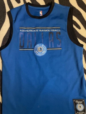Blå Dallas Mavericks basketlinne - Snyggt blått basketlinne med svarta kanter och tryck framtill med texten 'Mavericks Basketball Dallas' samt Dallas Mavericks-logga. Perfekt för basketträning eller streetwear. NBA-märke i nacken och patch nertill.
