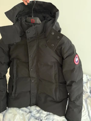 Canada Goose Wyndham  - Snygg svart Canada Goose Wyndham modell jacka med huva och klassisk logotyp på ärmen. använd några gånger bara nästan helt ny. Storlek S kan även vara som M. Jackan har dragkedja och knappar framtill, två stora fickor och detaljerade sömmar. Perfekt för kalla vinterdagar och riktigt varm.