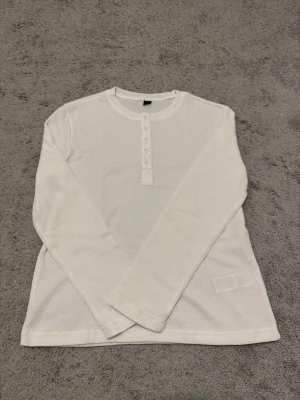 Vit långärmad henley tröja - Klassisk vit långärmad topp med rund halsringning och knappar framtill. Tillverkad i ett mjukt, ribbat material som ger en avslappnad och bekväm känsla. Perfekt att styla med jeans