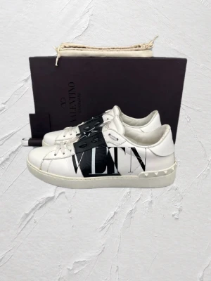 Valentino vita sneakers med svart detalj - Snygga vita sneakers från Valentino med svart band över snörningen, dekorerat med vita stjärnor och VLTN-logga på sidan. Skorna har en låg profil och vita nitar på hälen. Tillverkade i läder för en exklusiv känsla.