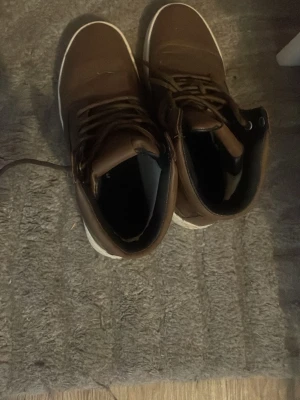 Bruna höga sneakers från Björn Borg - Snygga bruna kängor från Björn Borg med högt skaft och vita sulor. Skorna har snörning framtill och är gjorda i ett läderliknande material. Perfekta för dig som vill ha en stilren och urban look till hösten eller vintern.