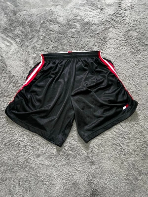 Vintage Tommy Hilfiger Mesh Shorts / Basketshorts - Snygga och klassiska vintage-shorts från Tommy Hilfiger i mesh-material. Modellen har en schysst retrostil med röd-vita revärer längs sidorna, vit resårmidja med dragsko samt den ikoniska logotypen på ena benet. Tillverkade i USA.