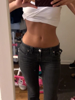 Svarta bootcut jeans från True Religion - Säljer ett par svarta bootcut jeans från True Religion med dekorativa sömmar och fickor med lock bak. Jeansen har låg midja, silverfärgade knappar och klassisk logga på bakfickan. Perfekta för en trendig och avslappnad stil.