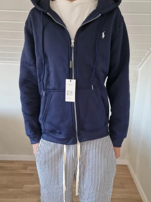 Ralph lauren zip hoodie - Snygg mörkblå hoodie från Ralph Lauren med dragkedja framtill och vit logga broderad på bröstet. Hoodien har huva med dragsko och två stora fickor framtill. Tillverkad i mjukt bomullsmaterial som känns skönt mot huden.