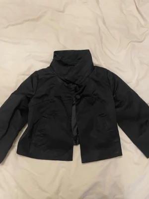 Svart kort kavaj med knappar - Snygg svart kort kavaj med hög krage och stora dekorativa knappar framtill. Kavajen har två fickor och är tillverkad i ett slätt, lätt glansigt material. Perfekt för dig som vill ha en stilren och trendig look.