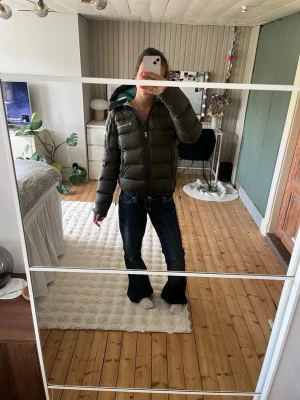 Mörkgrön dunjacka från Peak Performance - Snygg mörkgrön dunjacka från Peak Performance med luva och ljusblått foder. Jackan har dragkedja framtill och två sidofickor med dragkedja. Perfekt för kalla vinterdagar och har en modern, puffig design.