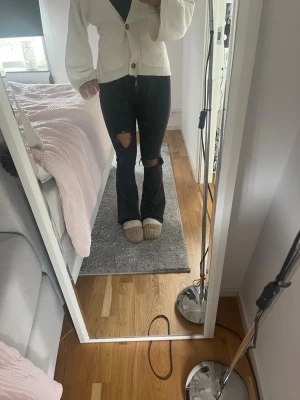 Svarta bootcut jeans med hål - Säljer ett par svarta bootcut jeans med slitna hål på båda knäna. Jeansen har hög midja och utsvängda ben, vilket ger en trendig och avslappnad look. Perfekta för dig som gillar en edgy stil och vill sticka ut lite extra.