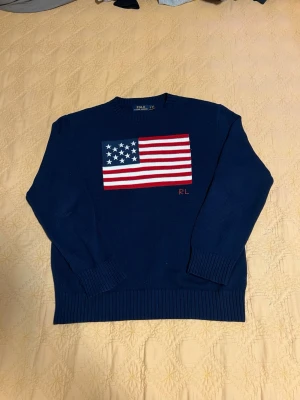 Mörkblå tröja Polo Ralph Lauren - Mörkblå stickad tröja från Polo Ralph Lauren med amerikansk flagga broderad på bröstet och RL-detalj. Tröjan har rund halsringning och ribbade muddar vid ärmslut och nederkant. Perfekt för dig som gillar klassisk och preppy stil.