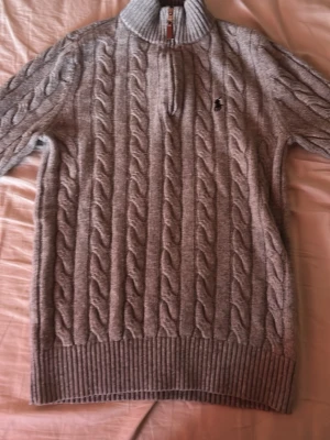Grå kabelstickad tröja med halvzip - Säljer en grå kabelstickad tröja med halv dragkedja och hög krage. Tröjan har ett diskret broderat märke på bröstet och breda ribbade muddar vid ärmslut och nederkant. Perfekt för lager på lager och riktigt snygg till jeans.