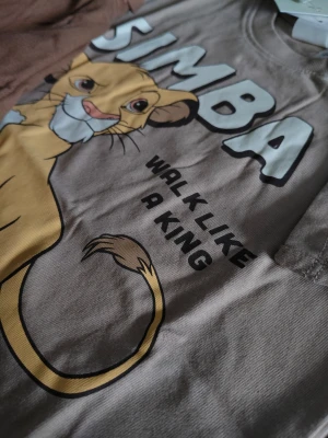 Beige Simba t-shirt med tryck med shorts - Shorts och T-shirt i beige med stort Simba-tryck framtill och texten 'WALK LIKE A KING'. T-shirten har rund hals och korta ärmar. Perfekt för dig som gillar Lejonkungen och vill ha en avslappnad stil. Obs Barnstorlek 74.