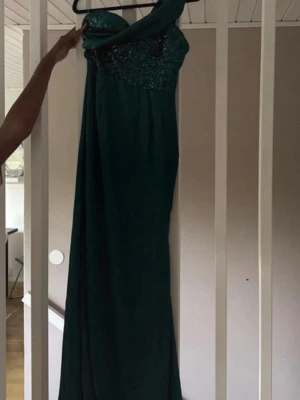 Grön långklänning med paljetter - Snygg grön långklänning med glittriga paljetter upptill och en axelbandslös design på ena sidan. Klänningen har oneshoulder och en elegant, figurnära passform som passar perfekt till speciella tillfällen.