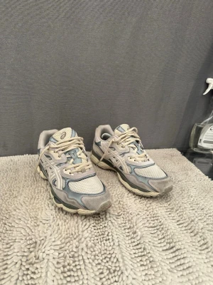 Asics gel NYC, Beige och grå sneakers med meshdetaljer - Snygga Asics sneakers i beige och grå toner med meshpaneler och detaljer i syntet. Skorna har en sportig look med rund tå, snörning och vadderad plös. Perfekta för dig som gillar en clean och stilren vibe.