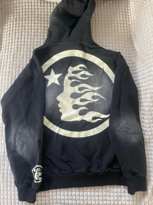 Svart hellstar hoodie med grafiskt tryck och en washed look - Säljer en svart hoodie med stor grafisk design på ryggen som föreställer en profil med eldflammor och en stjärna i en cirkel. Hoodien har huva och ribbade muddar. Perfekt för dig som gillar streetwear och unika prints.