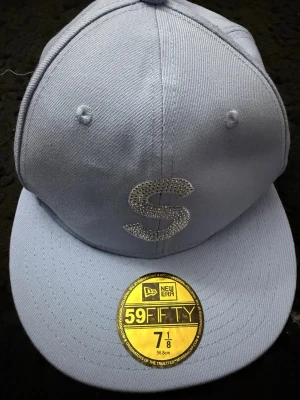 Ljusblå New Era keps - Snygg ljusblå keps från New Era, modell 59FIFTY. Kepsen har en rak skärm och ett stort S framtill i glittrande stenar. Tillverkad i bomull och polyester, perfekt för dig som vill sticka ut.