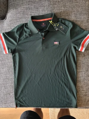 Mörkgrön piké med ränder - Snygg mörkgrön New Zeeland Auckland pikétröja med krage och knappslå. Ärmarna har vita och röda ränder för en sportig look. Liten patch med brittisk flagga på bröstet. Perfekt till jeans eller shorts.