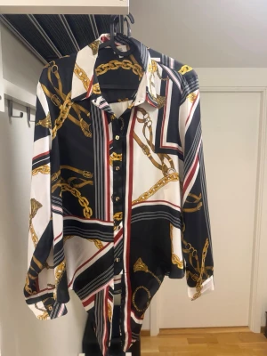 Mönstrad skjorta med kedjor och knyt - Snygg skjorta med svart, vit och röd bas, dekorerad med guldiga kedjemönster från River Island. Skjortan har långa ärmar, klassisk krage, knappar framtill och knyt i nederkanten. Materialet känns glansigt och liknar satin.
