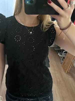 Svart blus med broderade hål - Säljer en svart blus från JAY med broderade hål i blommönster. Blusen har puffiga korta ärmar och rund halsringning.💕