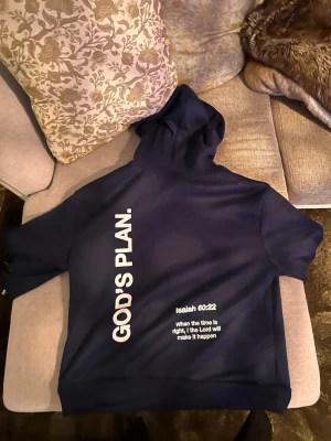 Mörkblå hoodie med texttryck - Snygg mörkblå hoodie med stor vit text 'GOD'S PLAN.' längs ryggen och bibelcitatet Isaiah 60:22 tryckt nedtill. Hoodien har klassisk huva och en avslappnad passform, perfekt för en streetwear-look.