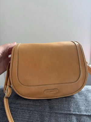 Ljusbrun axelväska i läderimitation - Snygg ljusbrun axelväska i läderimitation med rundade kanter och justerbar axelrem. Väskan har vita kontrastsömmar och enkel design utan synligt märke. Perfekt storlek för det viktigaste och stängs med lock. mått 25x20x10 cm 