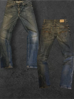 Blå slitna jeans från Nudie Jeans - Säljer ett par blå jeans från Nudie Jeans med slitningar och tvättade detaljer. Klassisk femficksmodell med raka ben och orange sömmar. Jeansen har en cool vintage-look och är gjorda i denim. Perfekta för dig som gillar en avslappnad och trendig stil.