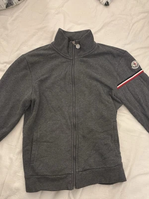 Grå zip hoodie från Moncler - Snygg grå zip hoodie från Moncler med broderad logga på ärmen och en röd, vit och blå randdetalj. Det är lite problem sturlit med dragkedjan men det går bra ändå.