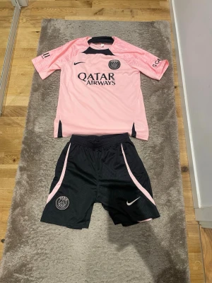 PSG set - Säljer ett PSG set från Nike med rosa tröja och svarta shorts. Tröjan har PSG-logga, Qatar Airways-tryck och svarta detaljer. Shortsen är svarta med rosa ränder och PSG-logga. Perfekt för fotbollsträning eller varma dagar
